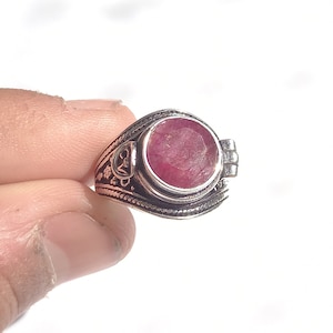 Può includere: Un anello d'argento con una pietra preziosa rossa e dettagli intricati. L'anello ha una parte superiore incernierata che si apre per rivelare un piccolo scomparto.