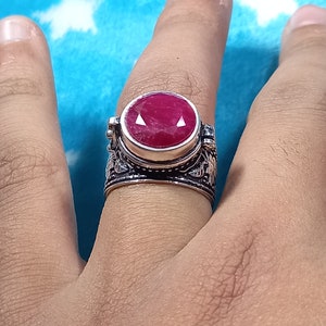 Puede incluir: Un anillo de plata con una piedra preciosa ovalada de color rojo intenso. El anillo tiene un diseño floral ornamentado alrededor de la banda y el engaste. La piedra preciosa es el punto focal, engastada en un bisel.
