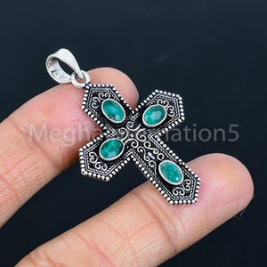 Pode incluir: Um pingente de cruz prateada com quatro pedras preciosas ovais verde-esmeralda. A cruz apresenta um design de filigrana ornamentado e uma argola na parte superior para uma corrente. O pingente é segurado pelos dedos de uma pessoa.