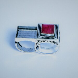 May include: Two silver rings with a rectangular design. One ring features a large, square, red gemstone. The other ring has an open design. The rings are set against a white background.