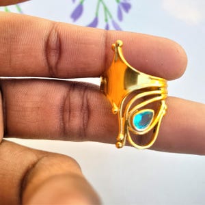 May include: A gold-colored ring with an ornate design, featuring a teardrop-shaped, light blue gemstone. The ring has a unique, openwork pattern with decorative elements and is held between fingers.