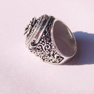 K&ouml;nnte beinhalten: Ein silberner Ring mit einem verzierten Design mit einem Scharnierdeckel und einem floralen Muster. Der Ring ist aus Metall und hat ein detailliertes Design.
