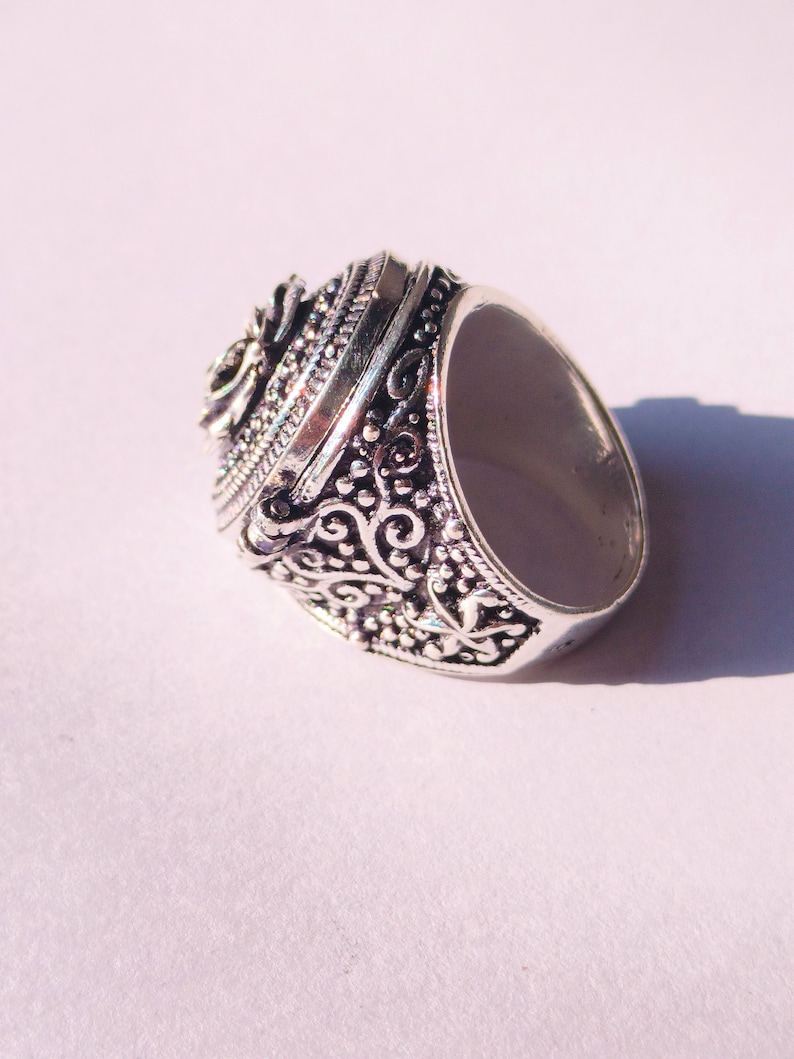 K&ouml;nnte beinhalten: Ein silberner Ring mit einem verzierten Design mit einem Scharnierdeckel und einem floralen Muster. Der Ring ist aus Metall und hat ein detailliertes Design.