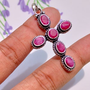 May include: A cross-shaped pendant featuring five oval-cut, deep pink ruby gemstones set in oxidized sterling silver. The silver setting has a twisted rope-like texture surrounding each stone, and a small silver bail is attached at the top.