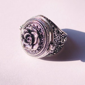 Könnte beinhalten: Ein silberner Ring mit einem detaillierten Rosenmuster auf der Oberseite. Der Ring hat einen Klappdeckel, der sich öffnet und ein kleines Fach freigibt.