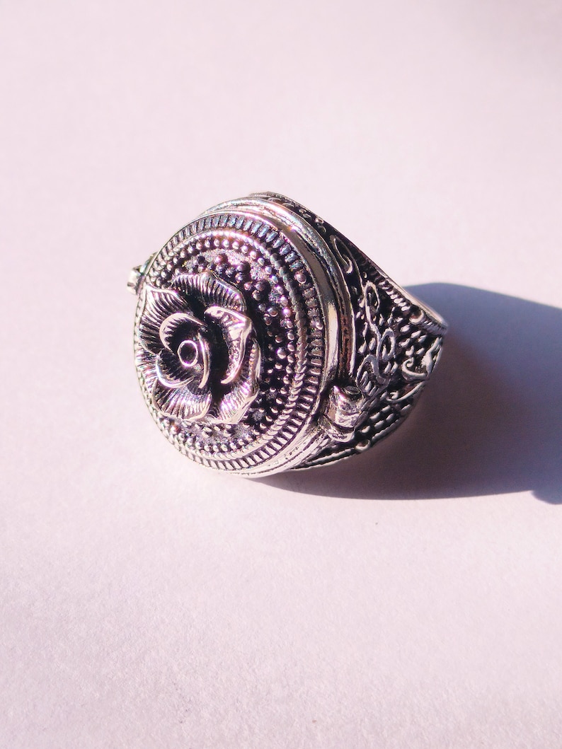 K&ouml;nnte beinhalten: Ein silberner Ring mit einem detaillierten Rosenmuster auf der Oberseite. Der Ring hat einen Klappdeckel, der sich &ouml;ffnet und ein kleines Fach freigibt.