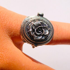 K&ouml;nnte beinhalten: Ein silberner Ring mit einem ovalen, klappbaren Anh&auml;nger mit einem detaillierten Rosenmotiv.