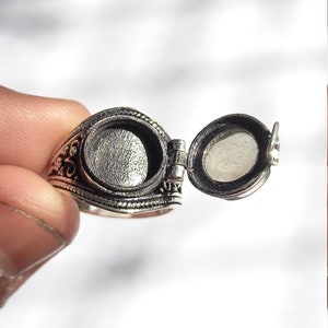 May include: A silver ring with a hinged, round locket that opens to reveal an empty space. The ring has an ornate design.