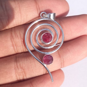 Puede incluir: Colgante de plata con diseño en espiral, con dos piedras preciosas redondas y rojas. El colgante tiene un lazo en la parte superior para una cadena. Las piedras preciosas son de color rojo intenso.