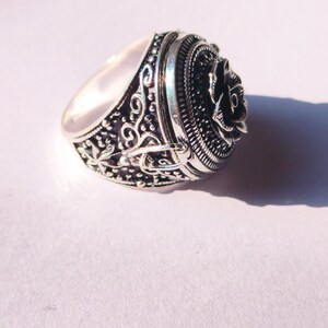 K&ouml;nnte beinhalten: Ein silberner Ring mit einem detaillierten Design, das eine Rose auf der Oberseite zeigt. Der Ring hat einen Klappdeckel, der sich &ouml;ffnet und ein kleines Fach freigibt.