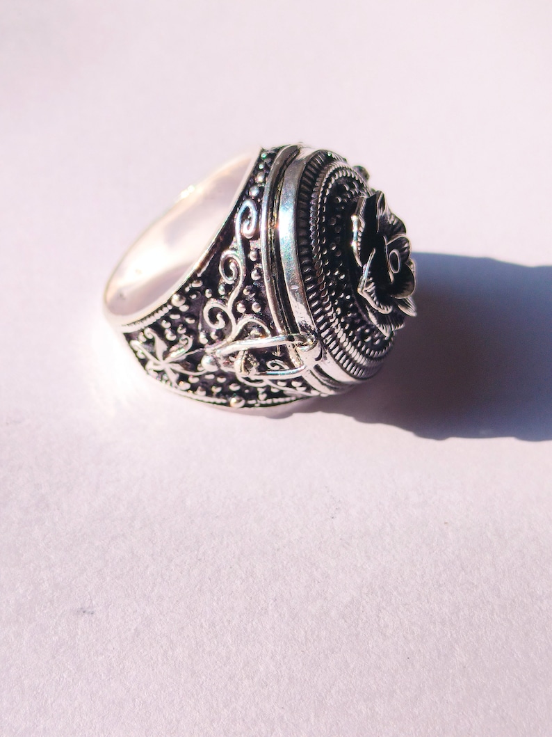 K&ouml;nnte beinhalten: Ein silberner Ring mit einem detaillierten Design, das eine Rose auf der Oberseite zeigt. Der Ring hat einen Klappdeckel, der sich &ouml;ffnet und ein kleines Fach freigibt.