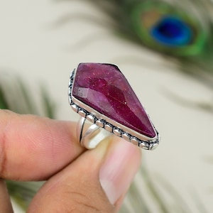 Może przedstawiać: Srebrny pierścionek z dużym, fasetowanym, głęboko czerwonym kamieniem szlachetnym. Kamień szlachetny ma wydłużony kształt z ostrym zakończeniem. Oprawa pierścionka ma dekoracyjną obwódkę z małych, okrągłych akcentów.