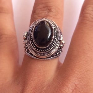 Puede incluir: Anillo plateado con una piedra negra ovalada y brillante. El anillo tiene un diseño detallado y ornamentado alrededor de la piedra y la banda. El anillo está en un dedo.