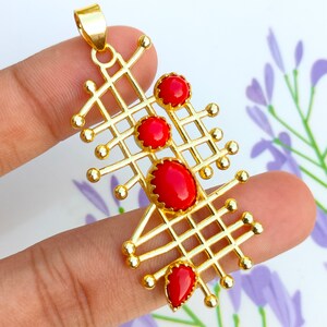 Puede incluir: Colgante dorado con diseño geométrico, con un patrón de rejilla y piedras rojas de forma ovalada y de lágrima. El colgante tiene un lazo en la parte superior para una cadena.