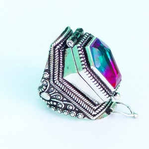 Op de afbeelding: Een zilverkleurige ring met een sierlijk, gedetailleerd ontwerp. De ring heeft een grote, veelzijdige edelsteen met een kleurengradatie van roze, paars, groen en blauw. De ring staat tegen een witte achtergrond.