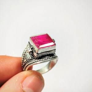 Pode incluir: Um anel de prata com uma gema de rubi rosa quadrada engastada em uma caixa decorativa com dobradiças. O anel é ornamentado e tem um design detalhado.