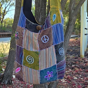May include: A patchwork shoulder bag with a black strap and a button closure. The bag features various fabric squares in shades of blue, green, brown, and purple, adorned with embroidered symbols like peace signs and flowers. The bag is hanging from a tree.