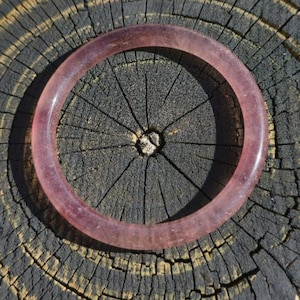 May include: A translucent, light purple bangle bracelet. The circular bracelet is made of a smooth, polished material. The bracelet is resting on a dark, textured wooden surface.