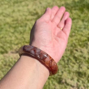 May include: A circular, polished bracelet with a mix of red and brown tones. The bangle is translucent and appears to be made of a natural stone. The bracelet is worn on a wrist, with a green grassy background.