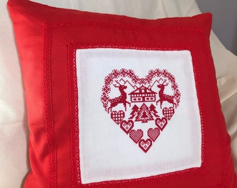 Puede incluir: Un cojín decorativo rojo con un centro blanco con un diseño de punto de cruz en forma de corazón. El diseño incluye ciervos, una casa, árboles y corazones. El cojín tiene un ribete de encaje.