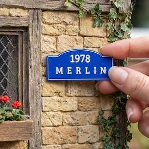 May include: A miniature blue street sign with white text reading "1978 MERLIN" is held in front of a detailed model of a brick building. The building has a window box with red flowers and climbing ivy.