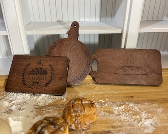 Puede incluir: Tres tablas de cortar de madera con temática de panadería. Una tabla tiene un logotipo con el texto "BAKERY" y una hogaza de pan. Otra tabla tiene una hogaza de pan con vapor saliendo de ella. La tercera tabla es lisa.