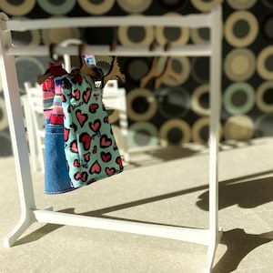 Puede incluir: Un perchero blanco con dos vestidos colgados. Los vestidos son un vestido a rayas azules y blancas y un vestido azul claro con corazones rosas.