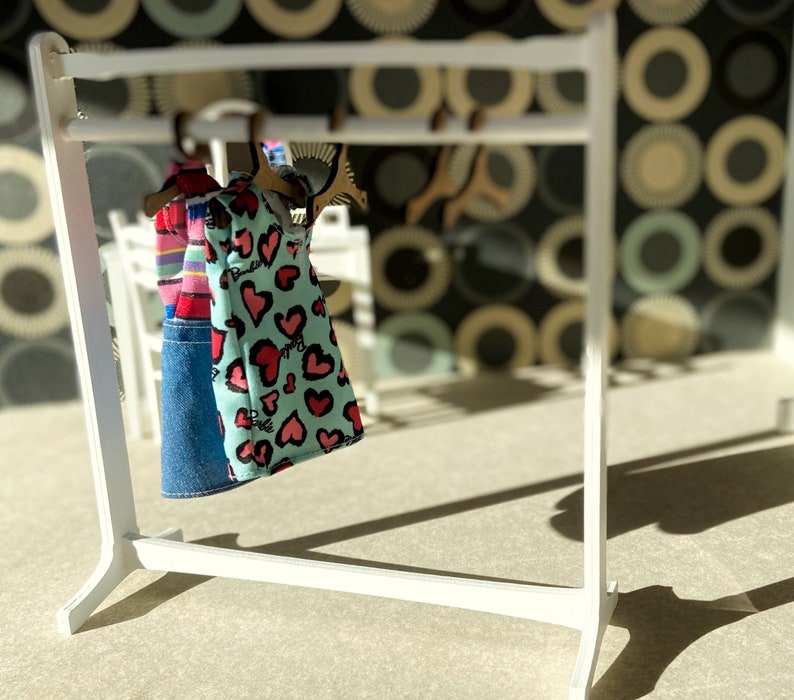 Puede incluir: Un perchero blanco con dos vestidos colgados. Los vestidos son un vestido a rayas azules y blancas y un vestido azul claro con corazones rosas.