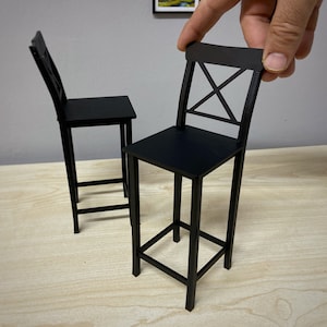 May include: Two black metal bar stools with a simple, modern design. The stools have a square base and a cross-shaped back.