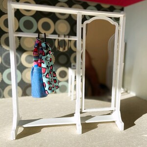 Peut inclure: Un portant &agrave; v&ecirc;tements en bois blanc avec un miroir et deux robes suspendues. Une robe est bleue &agrave; pois blancs et l'autre est rose &agrave; c&oelig;urs blancs.