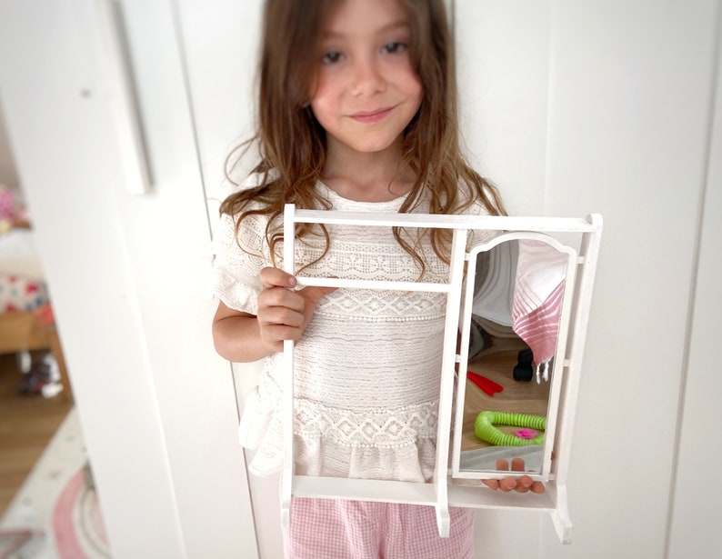 Peut inclure: Une coiffeuse jouet blanche avec un miroir et des &eacute;tag&egrave;res. La coiffeuse a une serviette ray&eacute;e rose et blanche accroch&eacute;e &agrave; l'arri&egrave;re. Un jouet vert et rose se trouve sur le sol devant la coiffeuse.