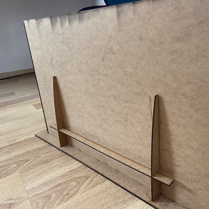 May include: A brown cardboard easel with two vertical supports and a horizontal base. The easel is designed to hold a sign or a piece of art.