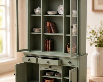 Könnte beinhalten: Ein salbeigrüner Miniatur-Buffetschrank mit verglasten oberen Türen, offenen Regalen sowie unteren Schubladen und Schränken. Die Regale enthalten kleine Dekorationsgegenstände wie Bücher, Teller, einen Krug und geflochtene Körbe.