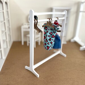 Puede incluir: Un perchero de madera blanco con una camisa estampada de color azul claro y rosa y una falda vaquera azul colgadas de perchas de madera marr&oacute;n.