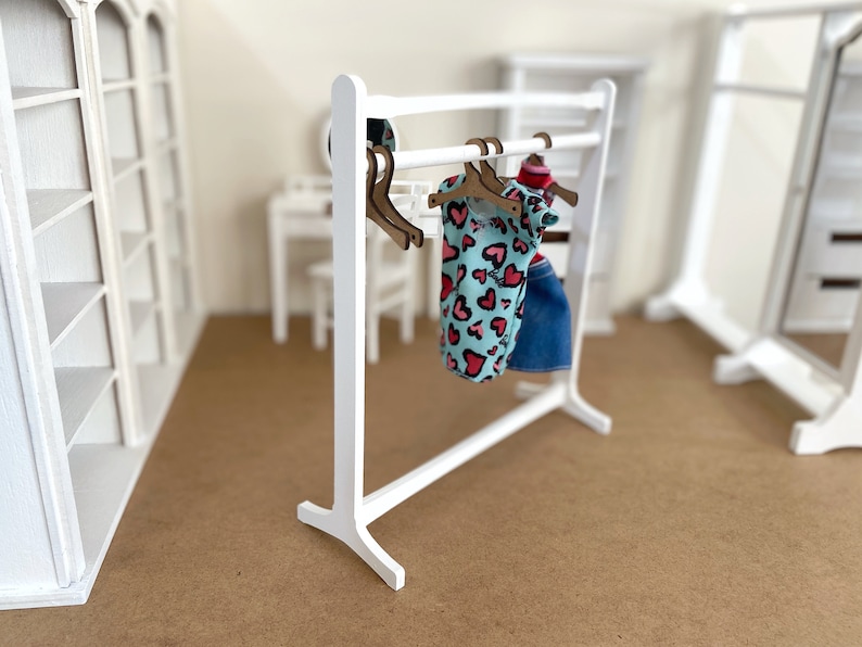 Puede incluir: Un perchero de madera blanco con una camisa estampada de color azul claro y rosa y una falda vaquera azul colgadas de perchas de madera marr&oacute;n.