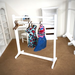Puede incluir: Un perchero de madera blanco con dos perchas que sostienen un vestido azul y blanco con corazones rosas y una falda vaquera azul con una camisa a rayas.