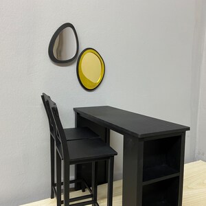 Pu&ograve; includere: Due sgabelli da bar neri con un tavolo nero con ripiani. Il tavolo ha un piano nero e due ripiani. Il tavolo &egrave; contro un muro bianco con due specchi decorativi, uno argentato e uno dorato.