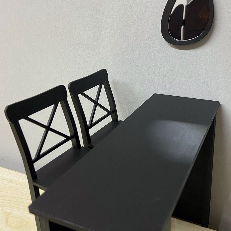 Pu&ograve; includere: Un tavolo nero con due sedie nere. Il tavolo ha un piano rettangolare e un design semplice. Le sedie hanno uno schienale a croce.