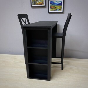 Pu&ograve; includere: Tavolo da bar in legno nero con uno scaffale e due sedie nere. Il tavolo ha un piano rettangolare e una struttura in metallo nero.