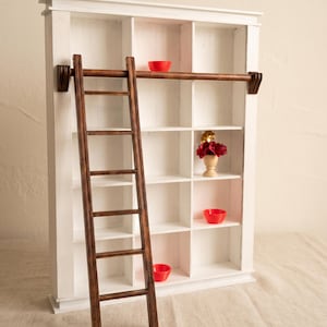 May include: A white wooden shelving unit with fifteen square compartments and a dark brown wooden ladder attached to a horizontal rail. Inside the shelves are three small red bowls and a miniature vase containing dark red flowers.