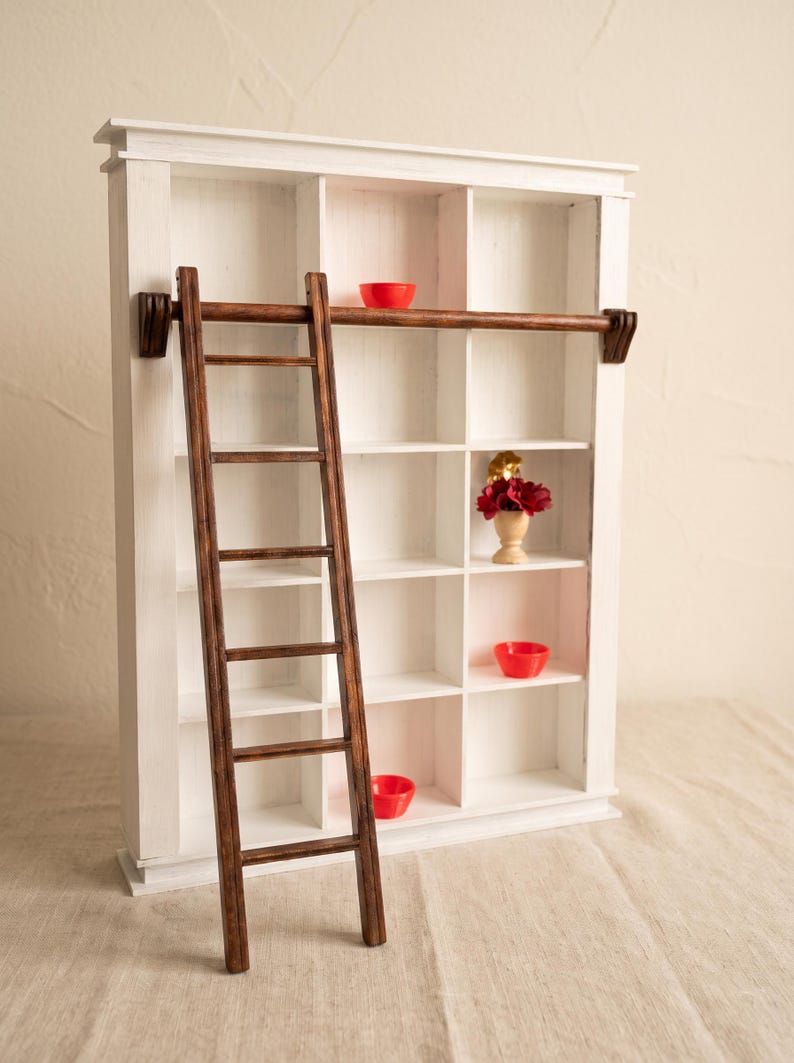 May include: A white wooden shelving unit with fifteen square compartments and a dark brown wooden ladder attached to a horizontal rail. Inside the shelves are three small red bowls and a miniature vase containing dark red flowers.