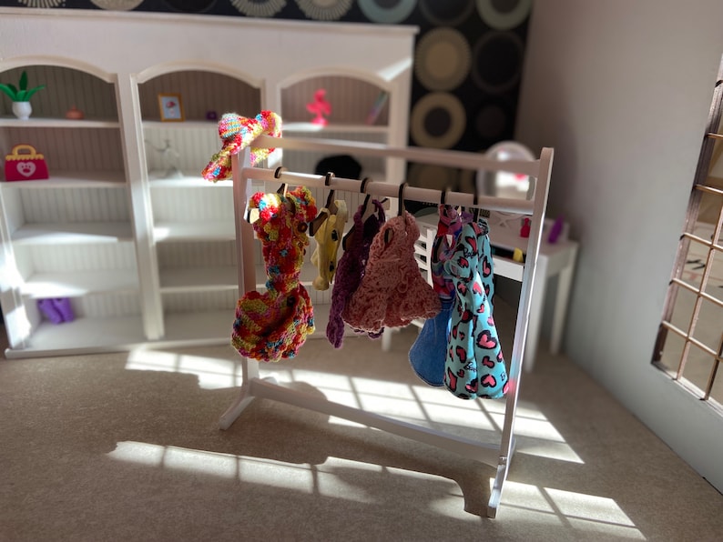 Puede incluir: Un perchero blanco con seis prendas de ropa colgadas. La ropa incluye un vestido de crochet rosa, un vestido de estampado de leopardo azul y rosa, un vestido de crochet amarillo y rojo, un su&eacute;ter de crochet morado, un vestido a rayas azul y blanco y un su&eacute;ter de crochet amarillo.