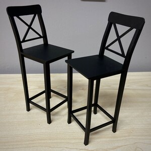 May include: Two black wooden bar stools with a simple, modern design. Each stool has a cross-shaped back and a square seat.