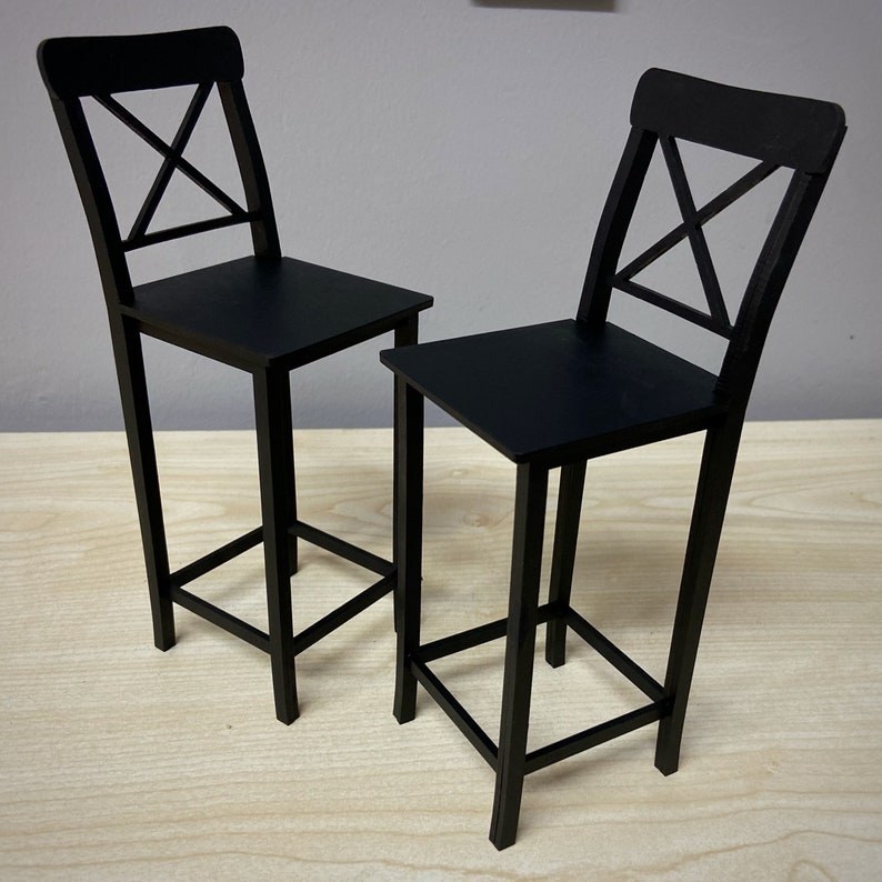 May include: Two black wooden bar stools with a simple, modern design. Each stool has a cross-shaped back and a square seat.