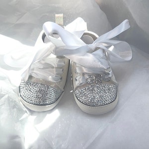 May include: A pair of white baby shoes with a white satin ribbon tied around them. The shoes are covered in rhinestones and have the word "GRO" spelled out in rhinestones on the front.