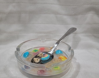 May include: A clear glass bowl filled with milk and colorful cereal, a silver spoon rests in the bowl.