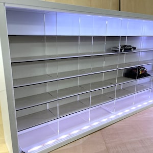May include: White display case with multiple shelves and clear dividers, illuminated by LED lights. A black car and a hat are visible on the shelves. The case is designed for displaying collectibles.