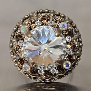 May include: A round, antique-style button with a large, faceted clear crystal center. The crystal is surrounded by smaller brown and clear crystals, set in a bronze-colored metal frame. The button has a detailed, ornate design.