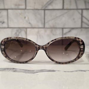 May include: Oval-shaped sunglasses with a brown gradient lens. The frames are a light brown color with a repeating diamond pattern. The sunglasses have a modern, stylish design.