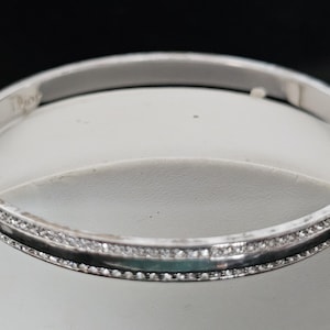 May include: A silver bangle bracelet with a row of small, clear gemstones set along the top and bottom edges. The bracelet has a smooth, polished finish and a simple, elegant design.